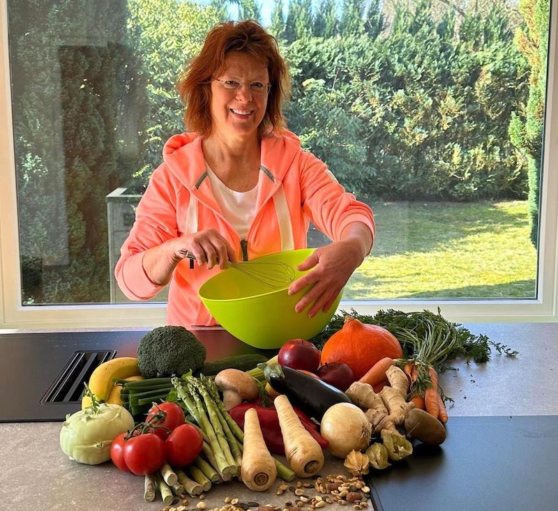 Antje Schlüschen mit Rührschüssel und frischem Gemüse auf der Arbeitsfläche vor sich; hinter ihr ein großes Fenster mit Blick in einen grünen Garten