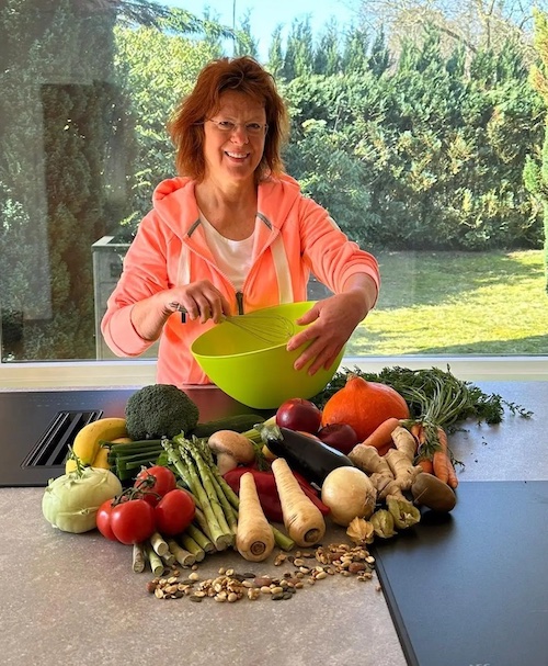 Antje Schlüschen mit Rührschüssel und frischem Gemüse auf der Arbeitsfläche vor sich; hinter ihr ein großes Fenster mit Blick in einen grünen Garten